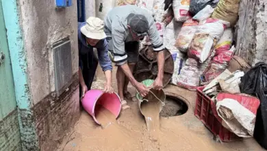 رجل بسيط يحمل دلوًا من الماء يزيل المياه المتراكمة من الطريق الضيق أمام منزل قديم. قام بتجميع بعض أكياس الرمل لحفظ الماء.